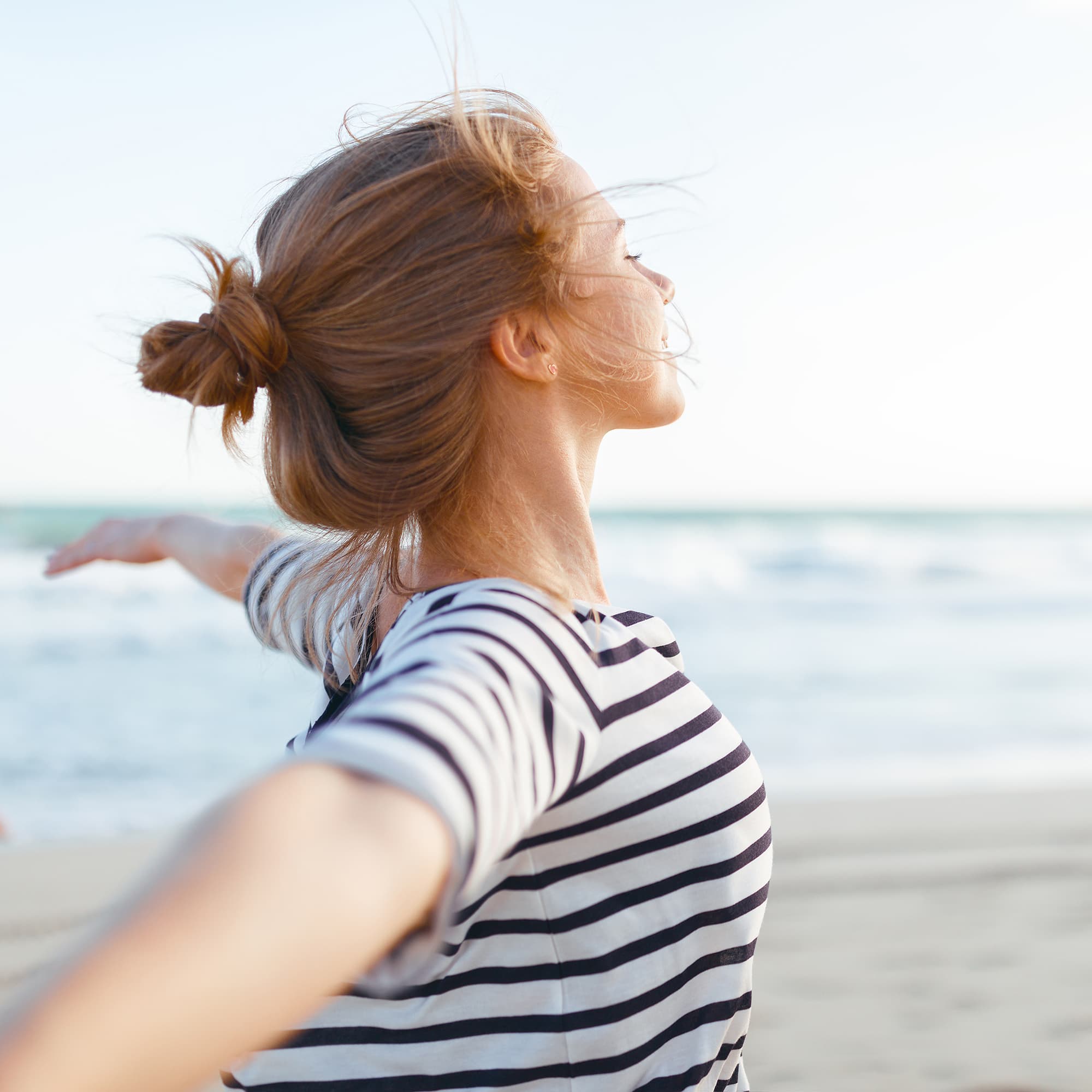 Frau mit gestreiftem Shirt genießt mit ausgebreiteten Armen den Wind am Strand