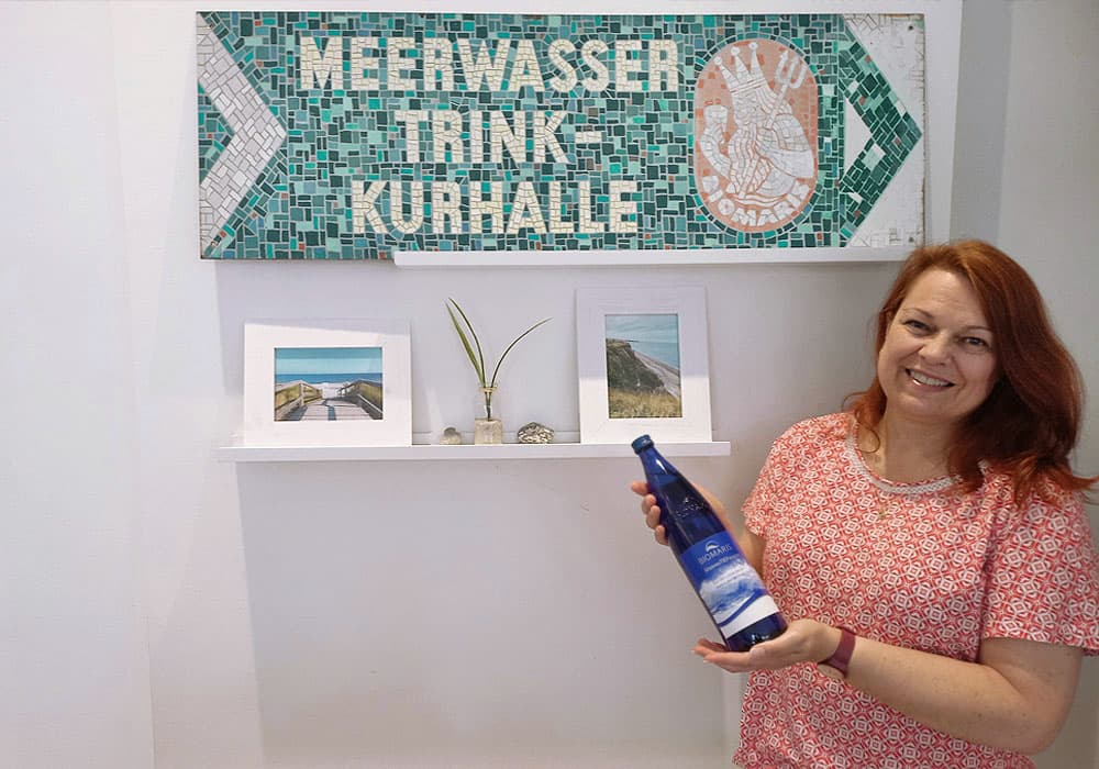 Shop-Betreiberin hält blaue BIOMARIS MeeresTIEFwasser Flasche vor Schild „Meerwasser Trink-Kurhalle“ , Bluse, Person, Portrait, Kräuter, Frau, Drinnen, Innenarchitektur, Werbung, Ärmel, Plakat
