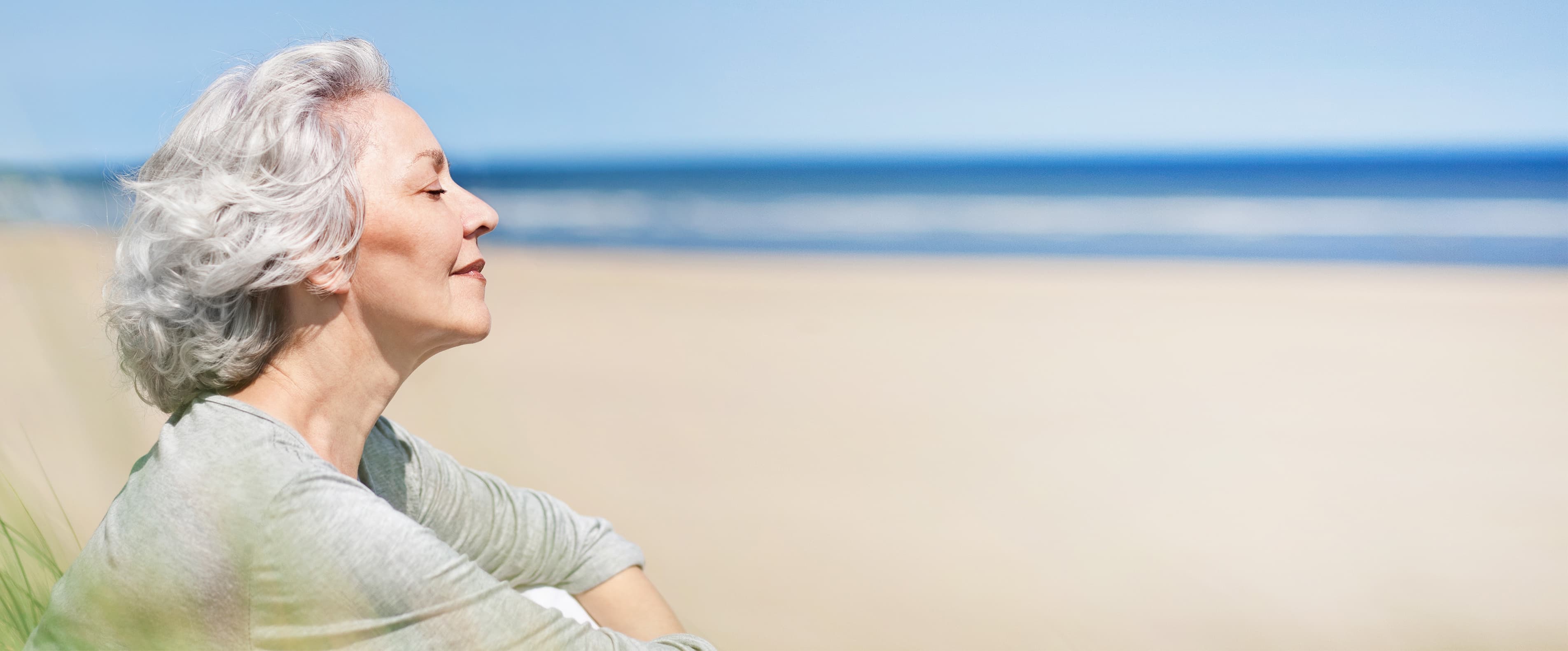 Am Strand Ältere Frau mit grauem Haar genießt mit geschlossenen Augen den Moment am ruhigen Sandstrand