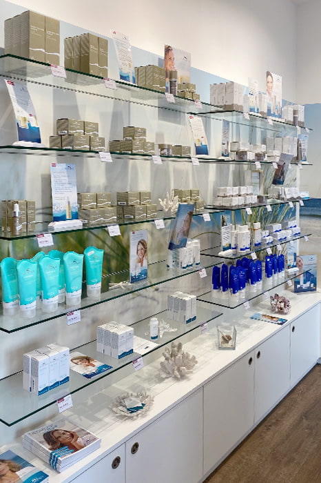 BIOMARIS Shop Scharbeutz Wandregal mit BIOMARIS Pflegeprodukten auf Glasböden, darunter weiße Unterschränke auf Holzfußboden