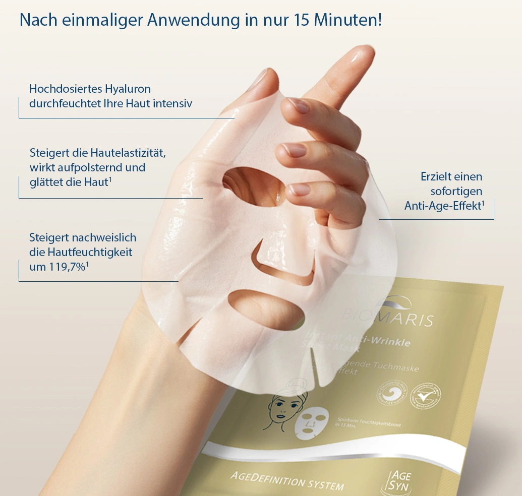 Hand hält eine weiße Gesichtsmaske mit Aussparungen für Augen, Nase und Mund, im Hintergrund die goldfarbene Verpackung.
