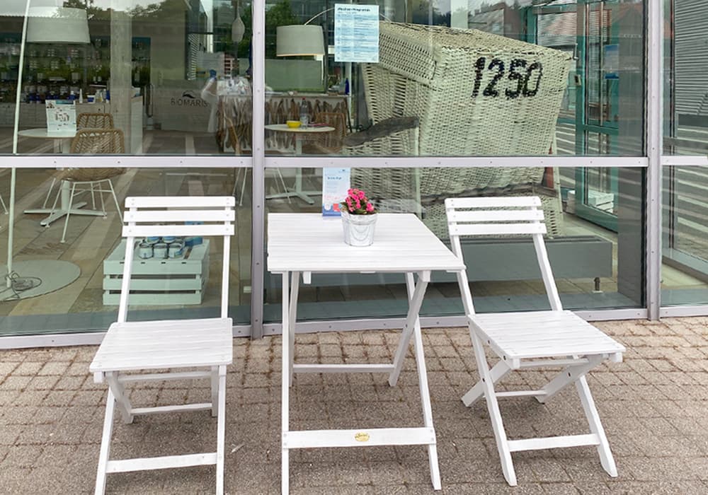 Weiße Sitzgruppe mit zwei Klappstühlen und Tisch mit Blumentopf vor Glasfassade mit Strandkorb im Hintergrund
