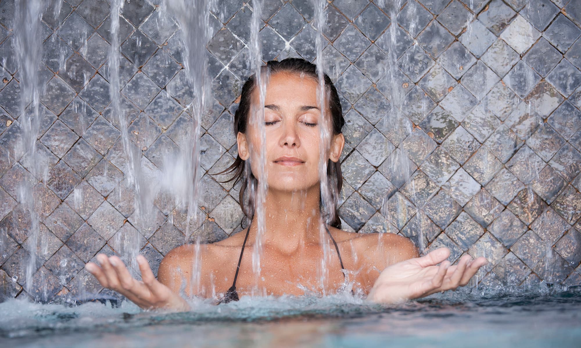 Spa Frau mit geschlossenen Augen genießt einen Wasserfall in einem Spa-Bereich vor gefliestem Hintergrund.