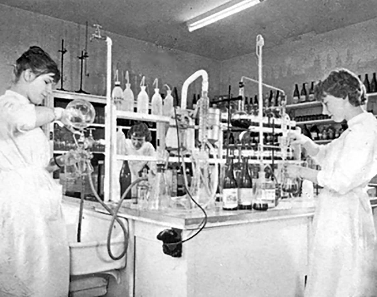 Drei Frauen in weißen Kitteln arbeiten konzentriert in einem Labor mit Glasgeräten und Reagenzflaschen.