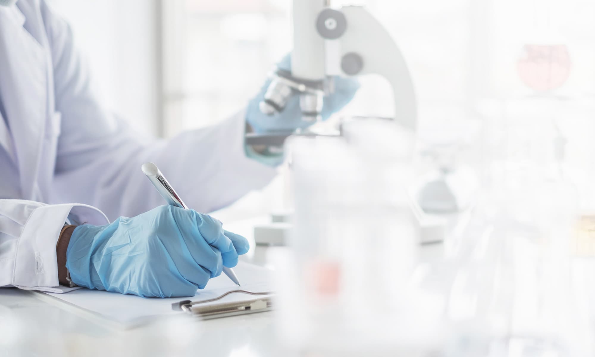 Forscher im Labor, schreibt mit blauen Handschuhen, Mikroskop im Hintergrund