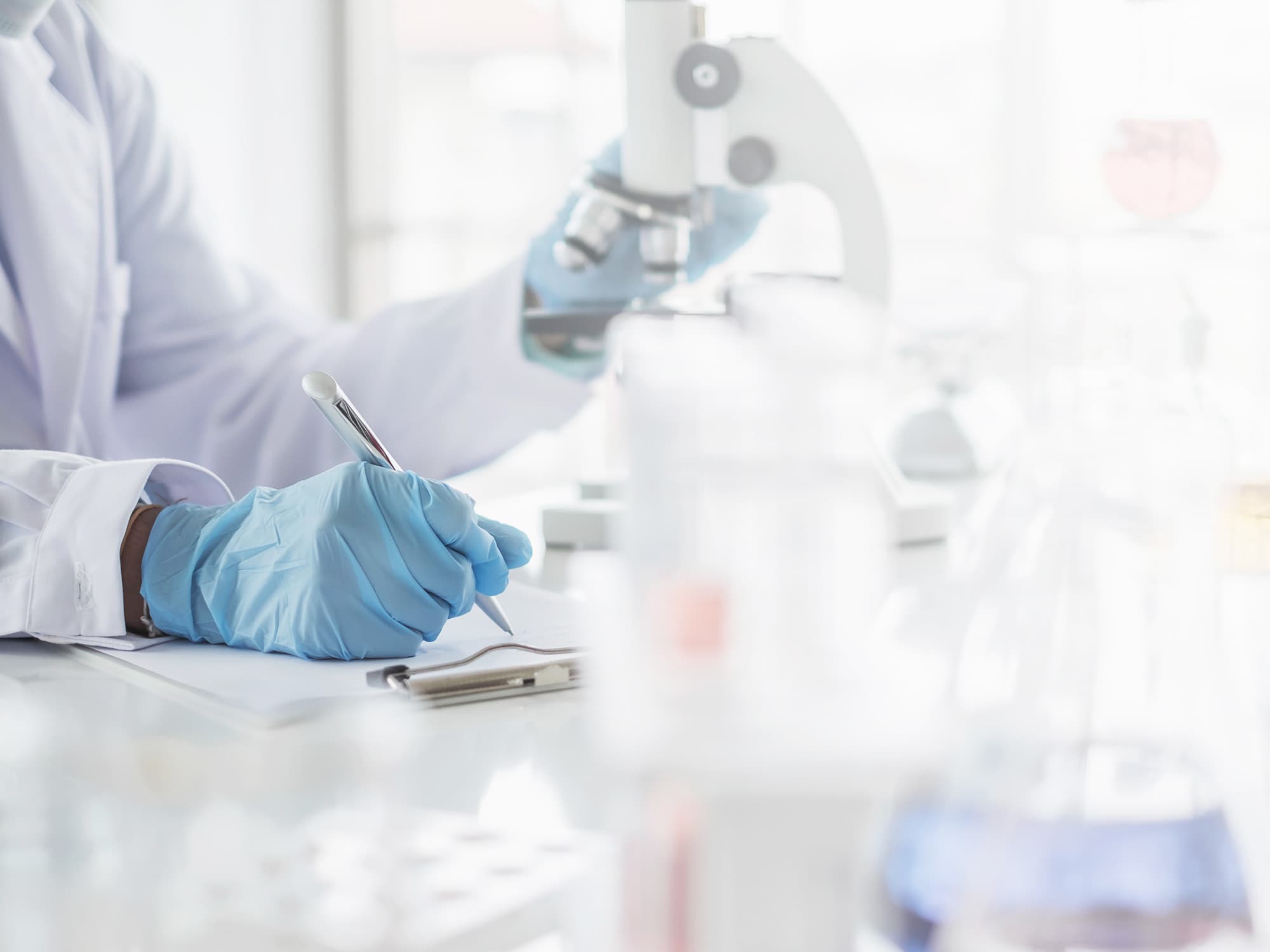 Laborarbeit Wissenschaftler mit blauen Handschuhen hält einen Stift und schreibt auf einem Klemmbrett, während er ein Mikroskop betrachtet.