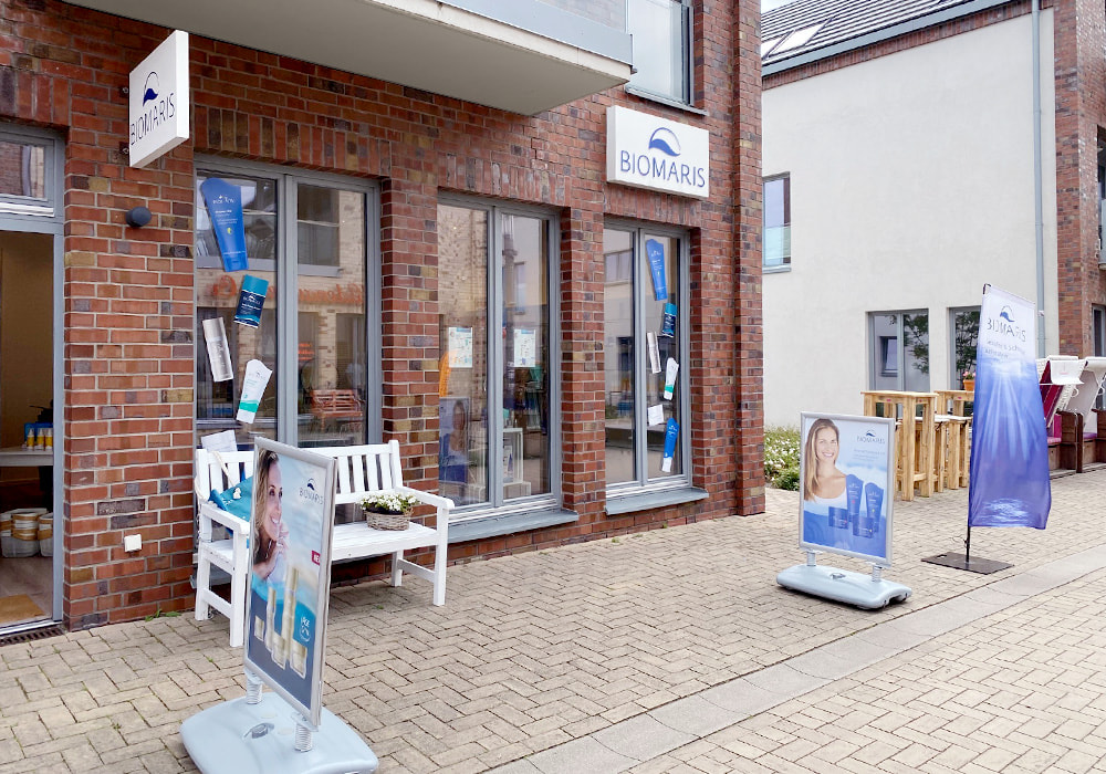 BIOMARIS Shop Heiligenhafen Außenansicht des BIOMARIS-Shops in Heiligenhafen mit roten Ziegeln, davor Werbetafeln, Fahne und Sitzbank