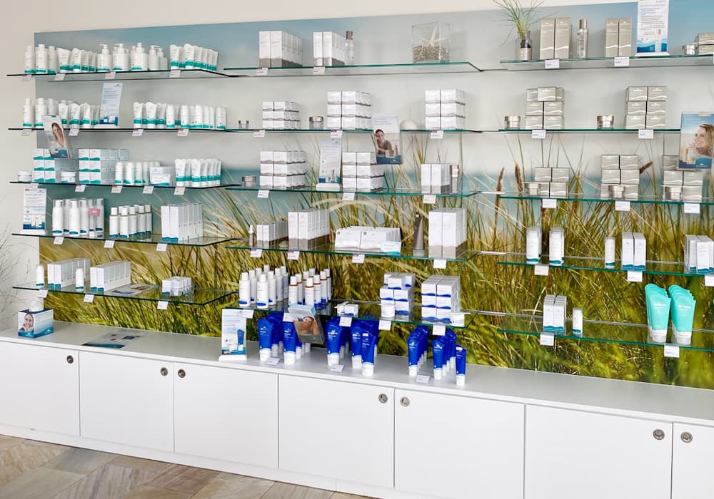 BIOMARIS Shop St. Peter-Ording BIOMARIS Verkaufsregal mit Pflegeprodukten auf Glasböden vor Wandbild mit Dünenlandschaft