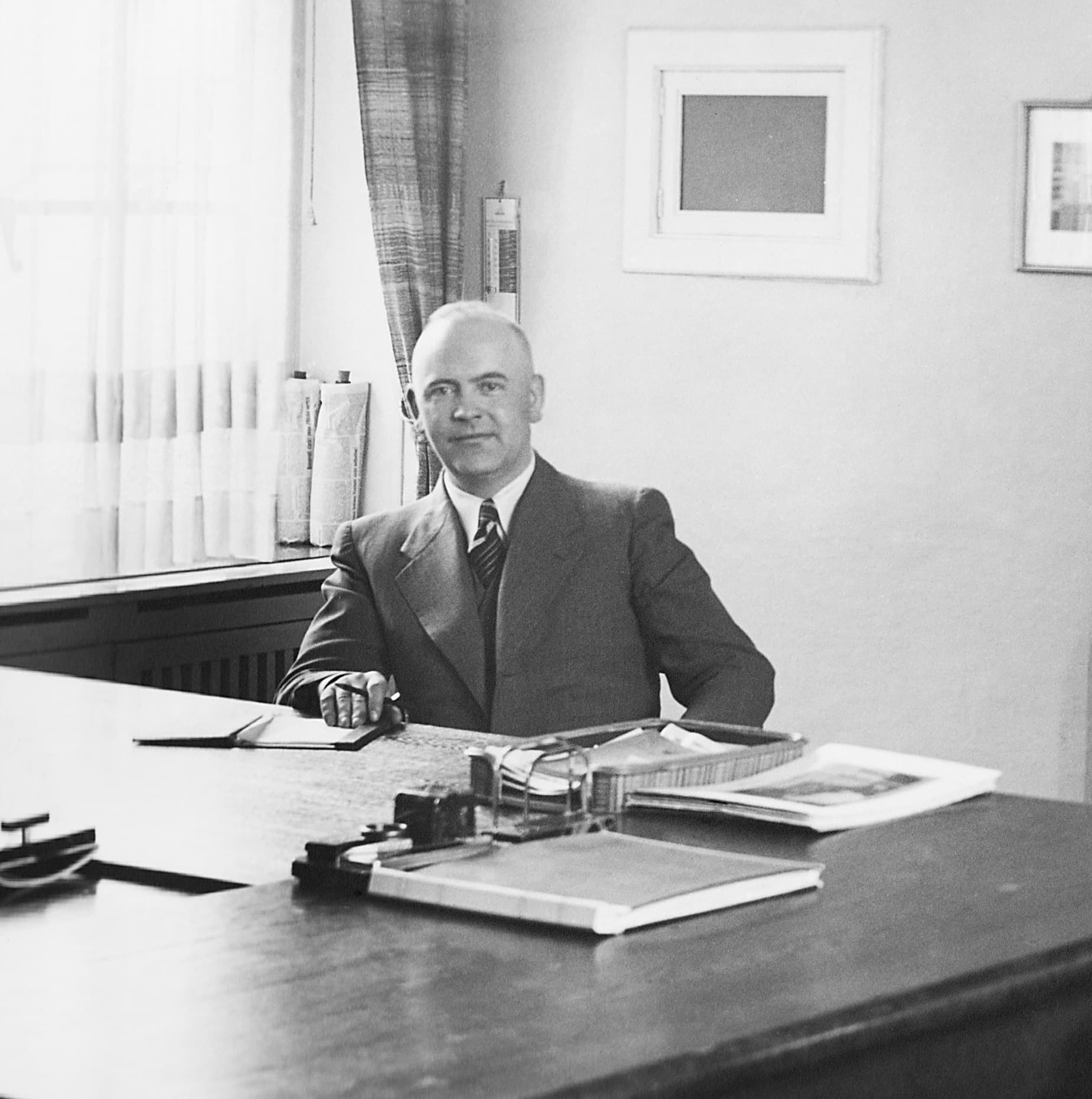 Schwarz-weiß-Porträt von Walter Brachmann, sitzend im Anzug an einem Schreibtisch in einem schlicht eingerichteten Büro.