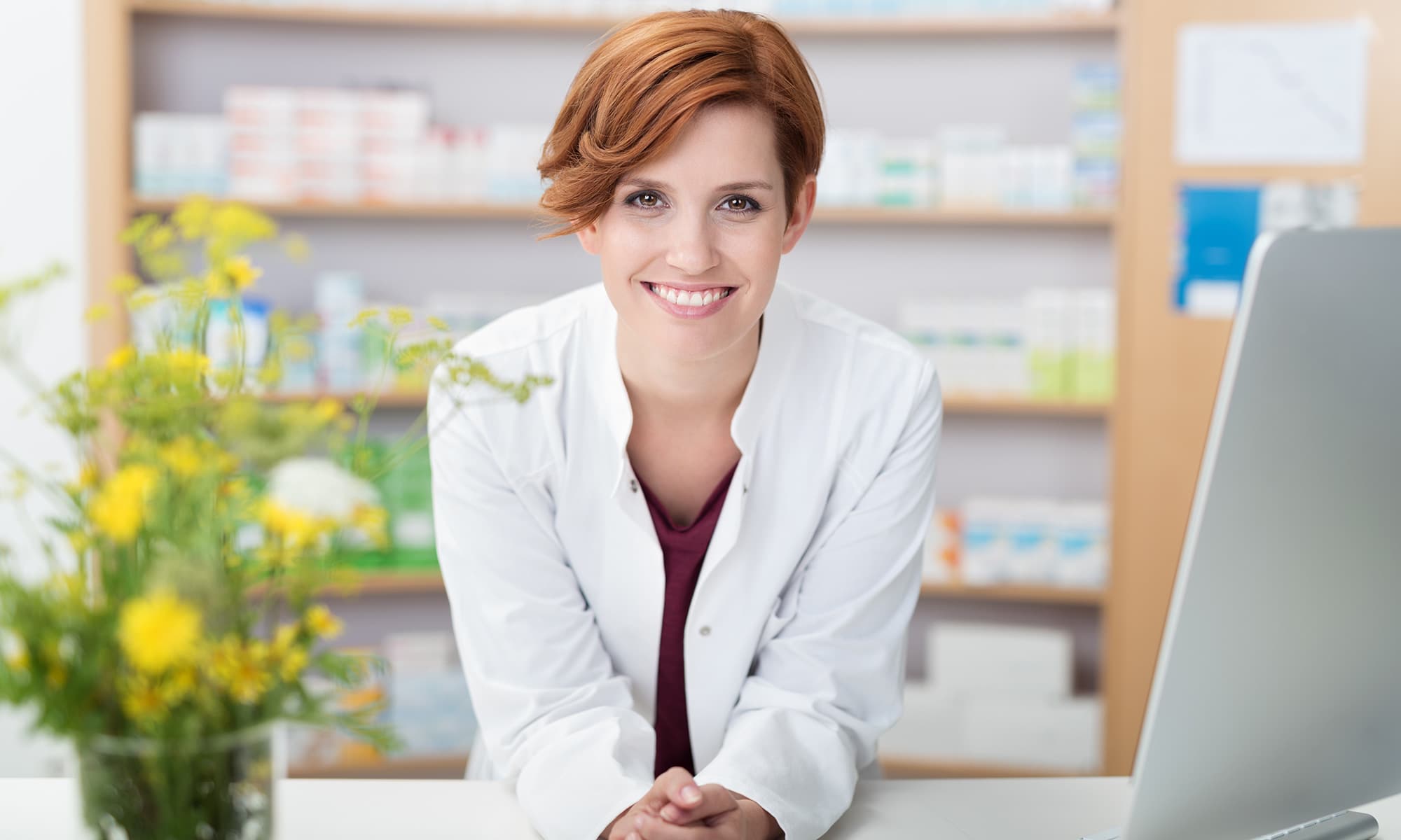 Apothekerin oder medizinische Fachkraft in weißem Kittel steht freundlich lächelnd vor einem Regal mit Medikamenten